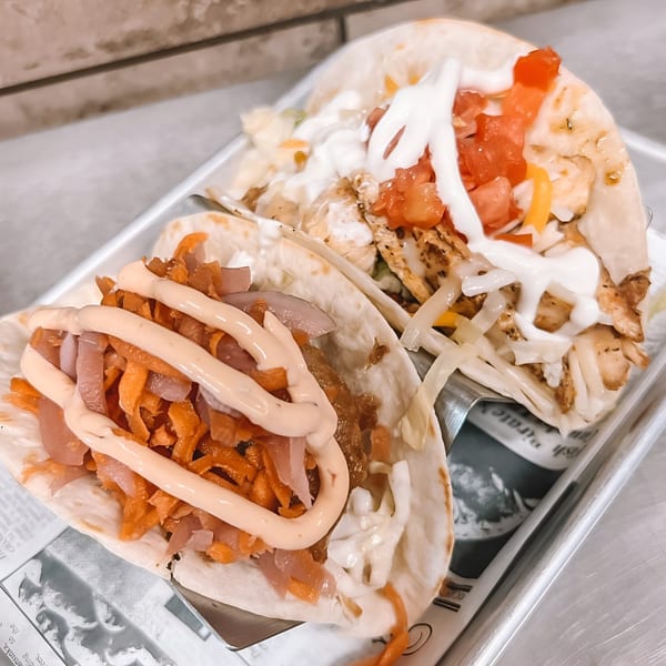 Two Jack And Harry's tacos, one with orange toppings and sauce, the other with white sauce and tomatoes.