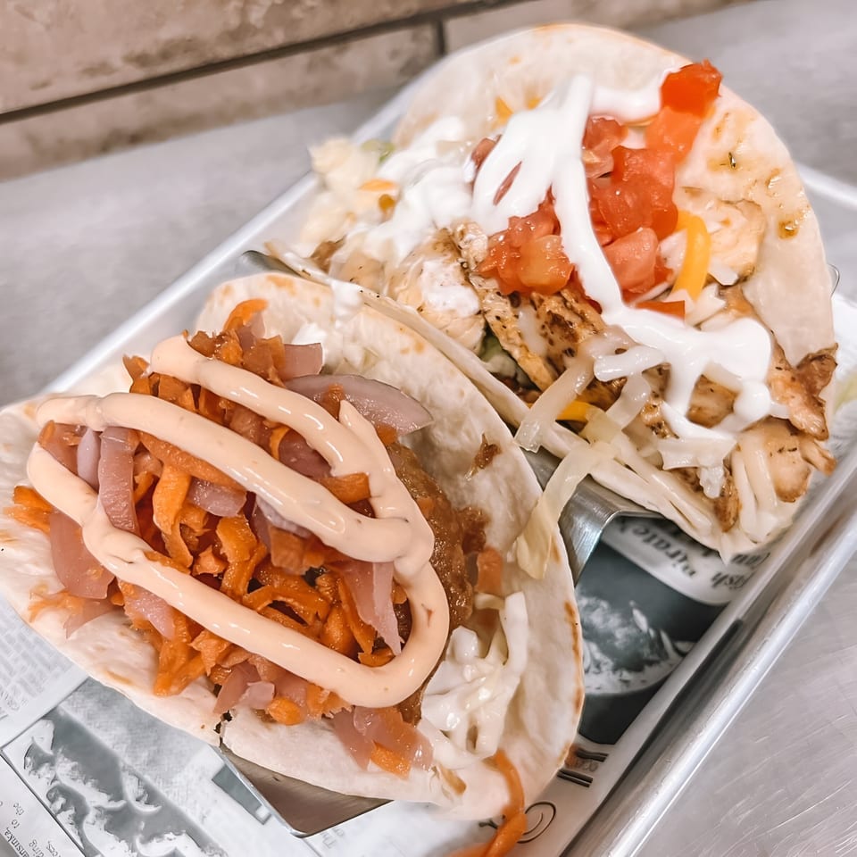 Two Jack And Harry's tacos, one with orange toppings and sauce, the other with white sauce and tomatoes.