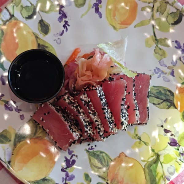 Seared tuna slices with sesame seeds, pickled ginger, and soy sauce on a lemon-patterned plate.
